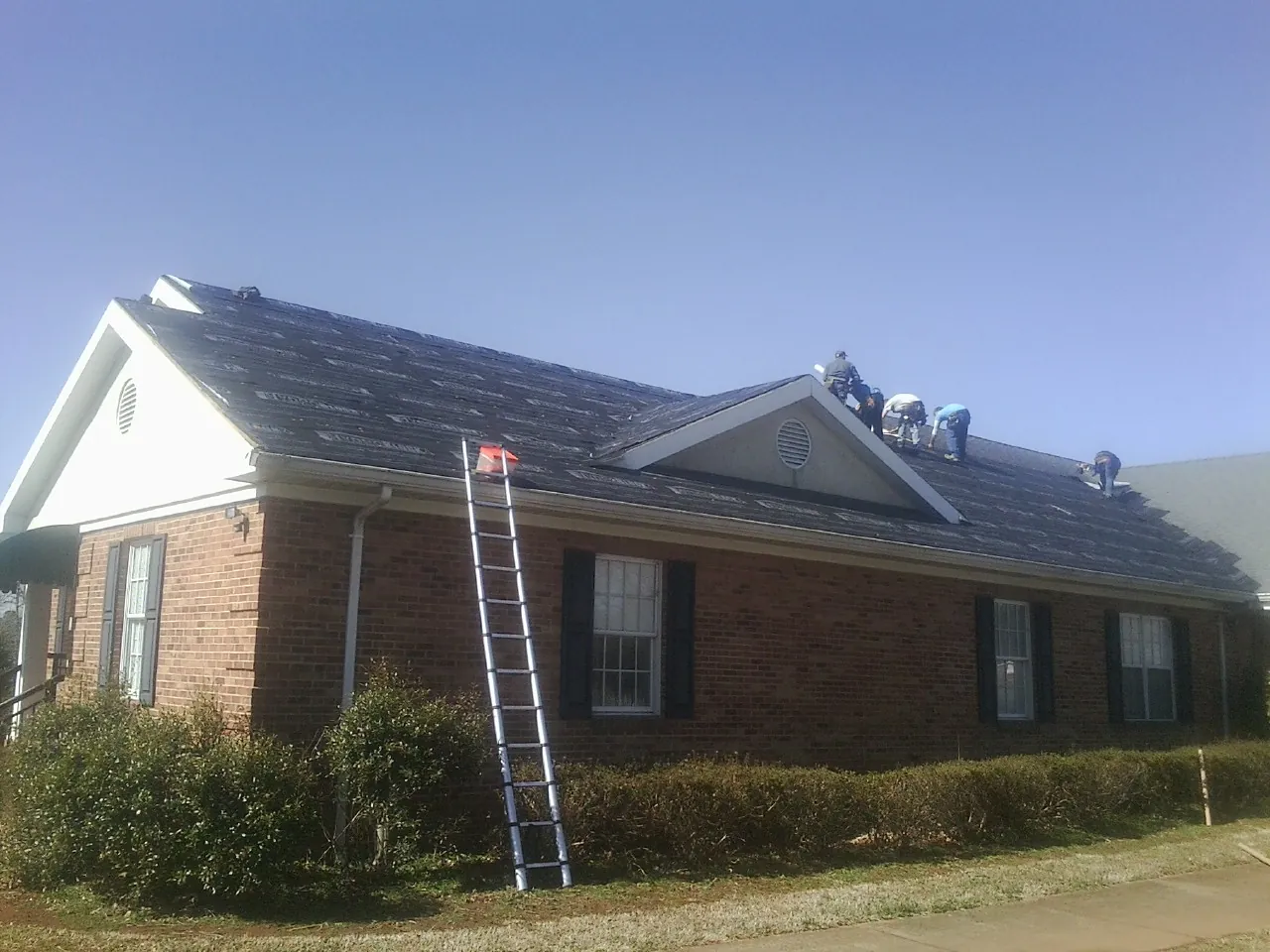 Crew tearing shingles off a brick ranch roof with a ladder set — Metal Roof Repair work by our crew in Southern Pines
