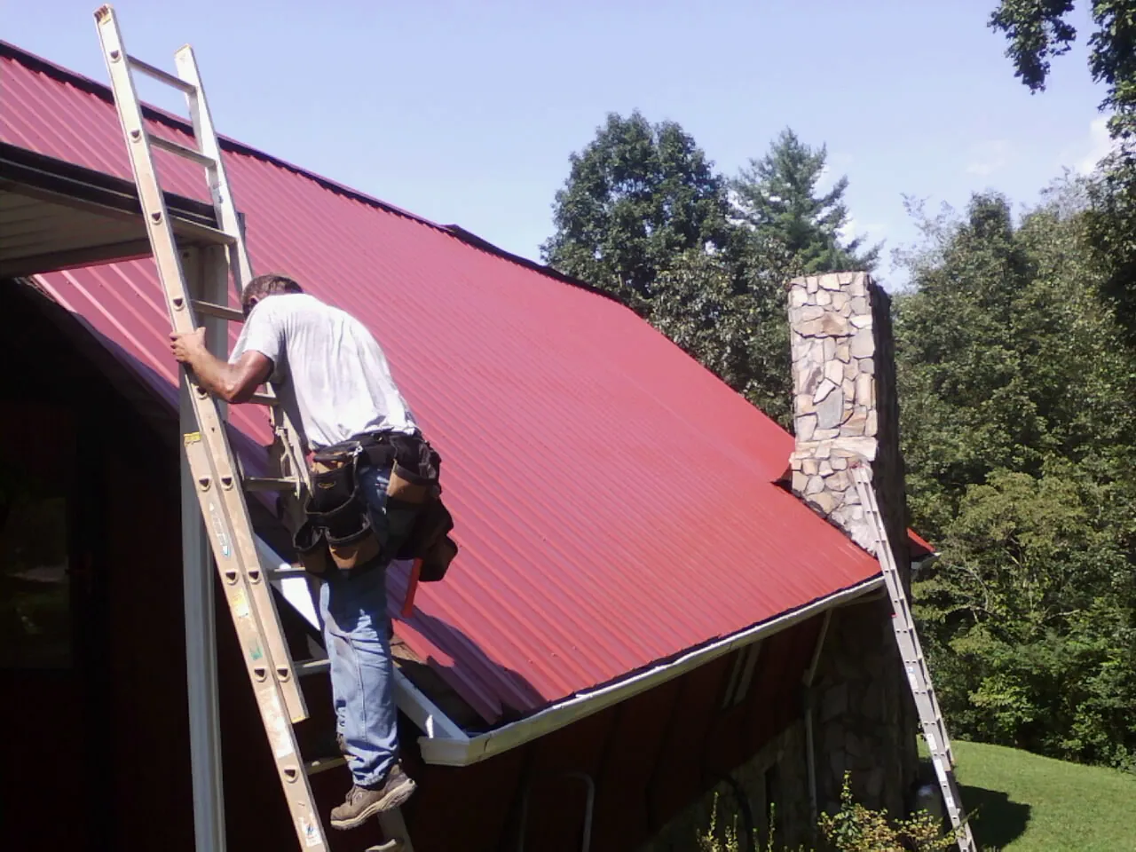 Roofer on a ladder beside a red metal roof and stone chimney — Soffit & Fascia Repair by Roofing Brothers in Southern Pines